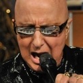 Close up of Paul Shaffer wearing a black sequin suit and singing into a microphone with his keyboard reflecting in his glasses