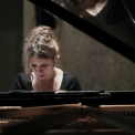 Image of Gabriela Monteri in a black shirt seated at a piano with eyes cast down toward the keys and her reflection visible in the lid of the piano.