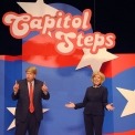 Two actors dressed as Donald Trump and Hillary Clinton debating one another in front of red white and blue background with Capitol Steps displayed overhead