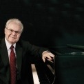 Emanuel Ax in a black suit sitting at his piano with a black background