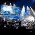 Band members playing on stage while white stage lights shine down on them and fake snow falls down. A screen behind the band projects a frozen landscape