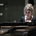 Image of Gabriela Monteri in a black shirt seated at a piano with eyes cast down toward the keys and the piano strings visible.