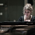 Image of Gabriela Monteri in a black shirt seated at a piano with eyes cast down toward the keys and the piano strings visible.