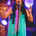 Woman in a turquoise dress and gold jewelry sings into a microphone with a glittery background