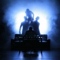 A black silhouette of a many sitting at a desk with three black silhouettes behind him in front of a black and blue smokey background.