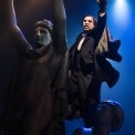 Phantom dressed in black with a white mask standing on two statues reaching out in front of a blue background.