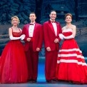 Two couples dressed in red standing arm in arm in front of a snowy cottage background