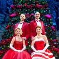 Two men standing behind two sitting women all dressed in red in front of a Christmas Tree