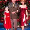 Man in uniform, woman in red dress, and young girl in red dress stand smiling in front of a Christmas Tree