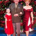 Man in uniform, woman in red dress, and young girl in red dress stand smiling in front of a Christmas Tree