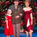 Man in uniform, woman in red dress, and young girl in red dress stand smiling in front of a Christmas Tree