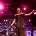 White woman, wearing a flowing black dress, singing into the microphone. Several pink lights in the background, as well as a purple fabric back-drop. Four other band members (trombone, violin, drums, and bass) are visible behind the singer.