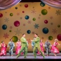 Two men in green shuts pointing at each other in front of a row of people in gray suits and red dresses dancing in front of a yellow background with ornaments and snowflakes on it