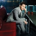 Michael Feinstein in a grey suit and tie and a white shirt sitting on a red leather couch in a dark room.