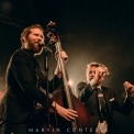 Two men wearing black suits, one with red hair and a beard and the other with curly brown hair. The red head is singing into the microphone and holding a stand-up bass. The man with brown hair is snapping his fingers.