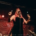 White women wearing a strapless black dress, has pink hair, and wearing many bead bracelets on her left hand, stands in the middle of the image singing into the microphone. Part of a drumset and trombone in the background of the image. Red fabric background.