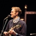 Chris Thile sings and plays mandolin wearing a collared shirt and sweater.