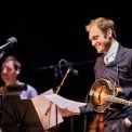 Chris Thile sings and plays mandolin wearing a collared shirt and sweater.