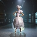 Woman in pink dress and pink hair in a room with a blue light behind her.