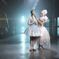 Woman in pink dress and pink hair with a woman in a blue dress and red shoes in a room with a blue light behind her