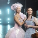 Woman in pink dress and pink hair with a woman in a blue dress and red shoes in a room with a blue light behind her