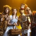 Man in lion costume, woman in blue dress and red shoes, man in tin man costume, and man in scarecrow costume standing next to each other in front of a yellow background.