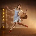 Woman in a blue dress and red shoes holding a basket jumping in front of a brown, lit up background.