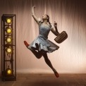 Woman in a blue dress and red shoes holding a basket jumping in front of a brown, lit up background.