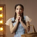 Woman in a blue dress and red shoes holding a basket in front of a brown, lit up background.