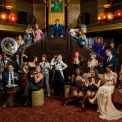 22 people in a 1920's styled room with brown, burgandy, and gold colors all variously standing and sitting. Some with instruments, some with whine glasses, all dressed in various 1920's costumes.