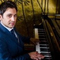 White man dressed in a blue suit with a blue and white striped tie, facing the camera as he plays the piano.