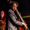 Man with black, curly hair, is wearing a grey suit and playing the bass instrument. White women with bright red hair, wearing a yellow and black dress, is standing behind the man.