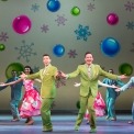 Two men in green suits dancing in front of a row of people in gray suits and red dresses dancing in front of a yellow background with ornaments and snowflakes on it