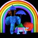 An imaginative animal puppet with an elephant head, a fox tail, a turtle back, and antlers wearing a hat and carrying an umbrella under a vibrant rainbow