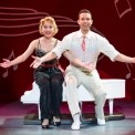 Man in white outfit and woman in black outfit sitting on a small white piano with arms presented forward