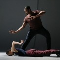 Man in a burgundy shirt and black pants standing and a man in a black shirt and burgundy pants laying down in front of him in front of a grey background.