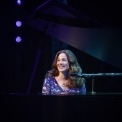 Woman in purple dress sitting behind a grand piano and microphone smiling