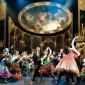 Dancers in masks and colorful gowns in front of a gold ballroom background