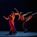 Two men dancing beside each other with a black background.  One man has his right arm curved up and his left arm curved down.  The other man has his back leg kicked up with his front arm lifted high.