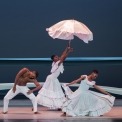 One male dancer wearing white squatting down and leaning to the side.  Two women dancers wearing white dresses. One woman is squatting and leaning to the side while holding her dress out.  The middle women has an umbrella that she is reaching to the sky. 
