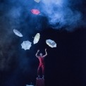 Man in red outfit standing on a red suitcase with seven umbrellas floating around him in front of a black and blue, smokey background.