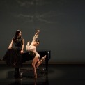 Women wearing a long black dress is sitting on the piano body facing another women who is dancing next to the piano and she dressed in white.