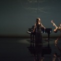 Woman wearing a long black dress standing in the middle of the frame, with a grand piano behind her. A women dressed in white dances to the women's left.