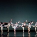 Nine women dressed in white dancing in front of a black background, with a piano and player in the back left corner