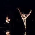 A man dressed in white dancing while facing the back, black background. Asian American woman playing on the piano in the background to the left.