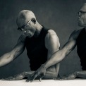 Two black and white images of the same man at a table in a black shirt and glasses in front of a grey background