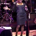 African American women, wearing a black sparkly shirt and a black skirt, is singing strongly into the microphone. Drummer wearing a yellow, white, and purple tie is playing in the background
