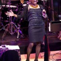 African American women, wearing a black sparkly shirt and a black skirt, is singing strongly into the microphone. Drummer wearing a yellow, white, and purple tie is playing in the background