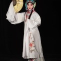 A Chinese woman wearing a white robe with flowers on it and a gold and blue headpiece stands holding a gold fan vertically near her face in front of a black background.