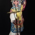 A Chinese woman wearing a headdress and robe made of ornate red, white, yellow, and blue fabric stands holding out a gold fan in front of a black background.